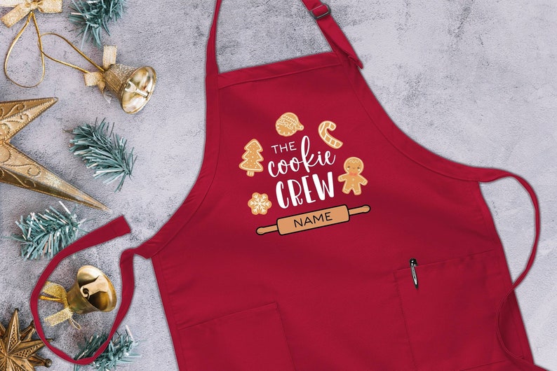 May include: Red Christmas cookie baking apron with a pocket and adjustable neck strap. The apron has a design that says "The Cookie Crew" with a gingerbread man, candy cane, snowflake, and rolling pin. There is a space for a name.