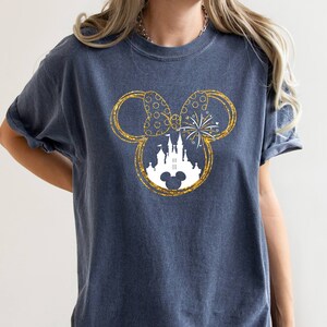 May include: A dark blue t-shirt with a gold glitter Mickey Mouse silhouette design. The silhouette includes a white castle and fireworks.