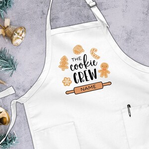 May include: White apron with a pocket and a festive design featuring gingerbread cookies, a rolling pin, and the text "The Cookie Crew".