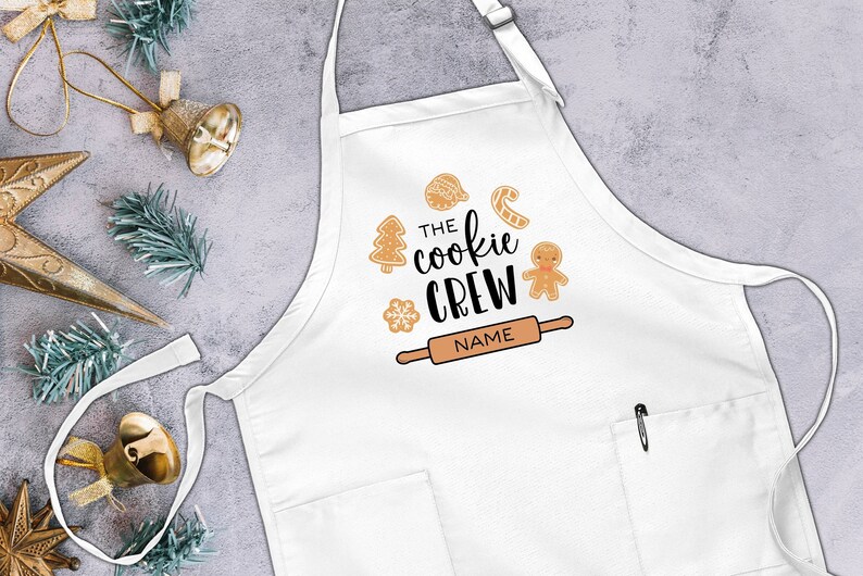 May include: White apron with a pocket and a festive design featuring gingerbread cookies, a rolling pin, and the text "The Cookie Crew".