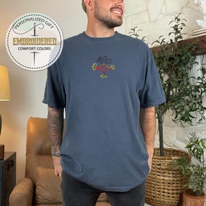 May include: A navy blue t-shirt with a yellow, red, and black embroidered Mickey Mouse design. The shirt has a relaxed fit and short sleeves.