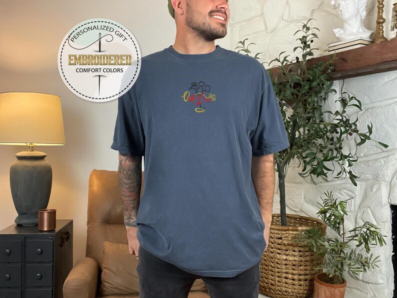 May include: A navy blue t-shirt with a yellow, red, and black embroidered Mickey Mouse design. The shirt has a relaxed fit and short sleeves.