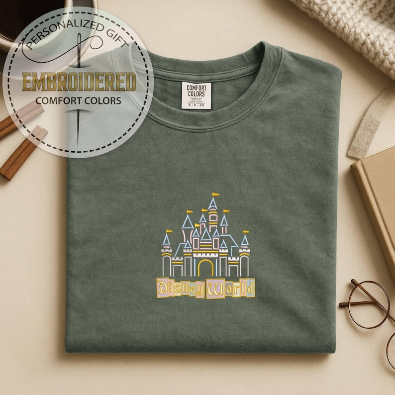 May include: A sage green t-shirt with an embroidered design of a castle with the text "Disney World" in yellow. The shirt has a crew neck and short sleeves. The words "Embroidered" and "Comfort Colors" are also visible.