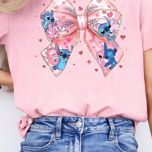 May include: Pink t-shirt with a large pink bow featuring a cartoon couple of Stitch and Angel. The bow is decorated with gold glitter and hearts.