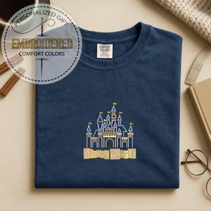 May include: Navy blue t-shirt with an embroidered Disney World castle design in white, yellow, and orange. The shirt features the text "Disney World" below the castle. The shirt also has a tag that says "Comfort Colors".