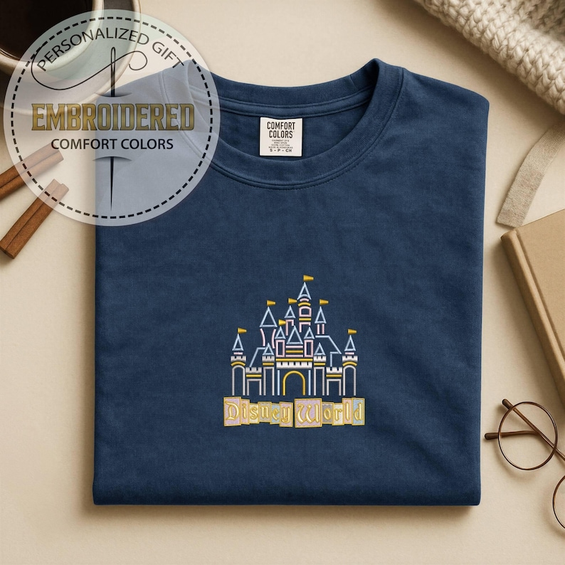May include: Navy blue t-shirt with an embroidered Disney World castle design in white, yellow, and orange. The shirt features the text "Disney World" below the castle. The shirt also has a tag that says "Comfort Colors".