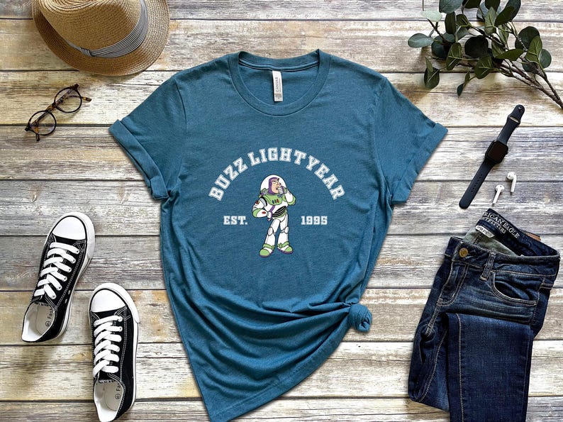 Może przedstawiać: Koszulka w kolorze turkusowym z białym nadrukiem przedstawiającym Buzz Lightyeara z serii Toy Story. Nadruk zawiera tekst "Buzz Lightyear" i "Est. 1995".