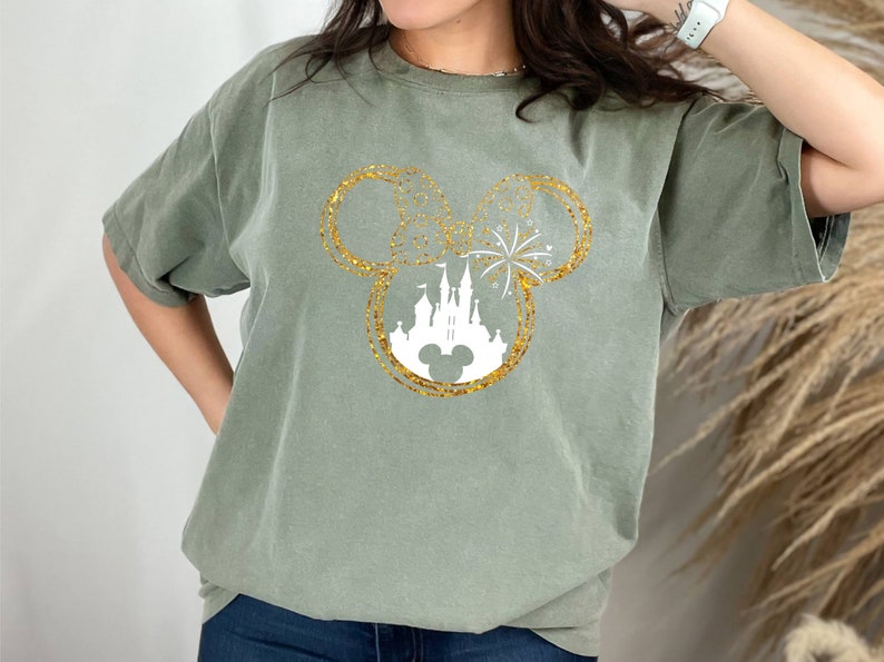 May include: A green t-shirt with a gold glitter Mickey Mouse head design featuring a white silhouette of Cinderella's castle and fireworks.