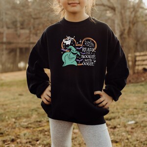 Pu&ograve; includere: Felpa nera con una grafica di Oogie Boogie, un fantasma e pipistrelli, con la scritta "Ready to Boogie with Oogie!". Il design &egrave; nei toni del verde, bianco e arancione. La felpa &egrave; indossata da una persona.