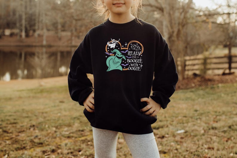 Pu&ograve; includere: Felpa nera con una grafica di Oogie Boogie, un fantasma e pipistrelli, con la scritta "Ready to Boogie with Oogie!". Il design &egrave; nei toni del verde, bianco e arancione. La felpa &egrave; indossata da una persona.