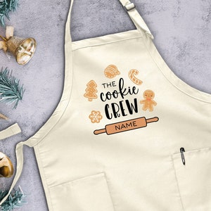May include: A white cotton apron with a pocket and adjustable neck strap. The apron has a festive design with the text "The Cookie Crew" and a gingerbread man, candy cane, and rolling pin.