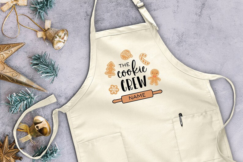 May include: A white cotton apron with a pocket and adjustable neck strap. The apron has a festive design with the text "The Cookie Crew" and a gingerbread man, candy cane, and rolling pin.