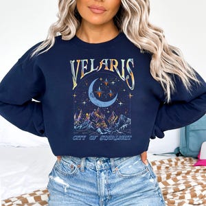 May include: Navy blue sweatshirt featuring the word "VELARIS" in a decorative font above a crescent moon and stars design. Below the moon is a mountain range with colorful peaks, and the text "CITY OF STARLIGHT".