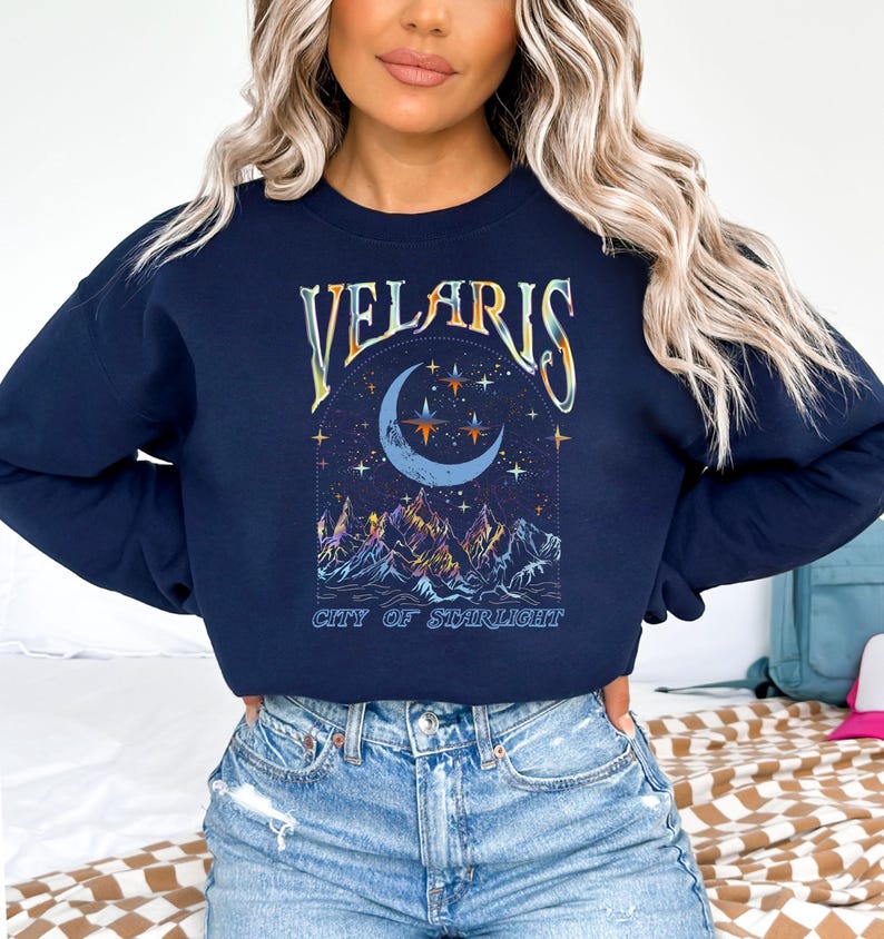 May include: Navy blue sweatshirt featuring the word "VELARIS" in a decorative font above a crescent moon and stars design. Below the moon is a mountain range with colorful peaks, and the text "CITY OF STARLIGHT".