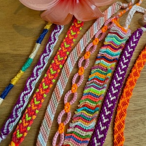 May include: A collection of colorful handmade friendship bracelets in various patterns and designs. The bracelets feature vibrant hues of orange, pink, purple, green, and blue, with intricate woven and knotted details. A translucent pink flower is in the background.