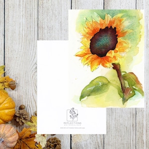May include: Watercolor painting of a sunflower with yellow and orange petals and a dark brown center. The painting is on a white card, with a blank card next to it. The background is a rustic wooden surface with pumpkins and leaves.