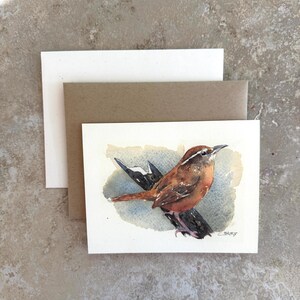 May include: A rectangular greeting card featuring a watercolor painting of a brown bird with a white eyebrow stripe perched on a dark branch against a blue wash background, accompanied by a white envelope and a brown kraft paper envelope.