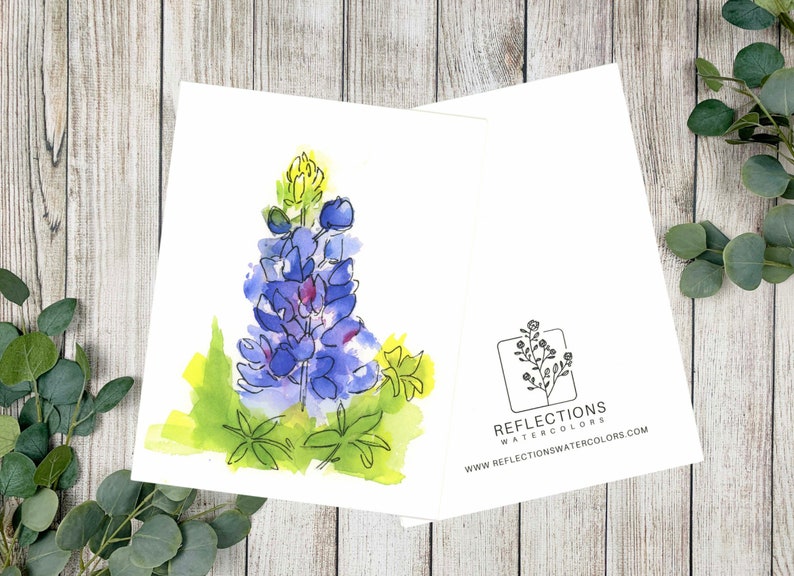May include: Open greeting card with a watercolour bluebonnet flower illustration. The card shows a detailed image of the flower in shades of purple and blue, with green and yellow accents. The back of the card has a logo and website address.