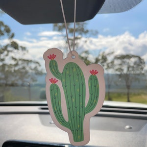 May include: A white car air freshener shaped like a green cactus with red flowers. The cactus is hanging from the rearview mirror of a car.