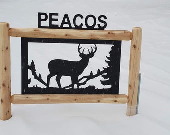 Op de afbeelding: Een houten frame met een metalen silhouet van een hert erin. Het frame staat in de sneeuw en het metalen silhouet is zwart. De naam "PEACOS" staat bovenop het frame.