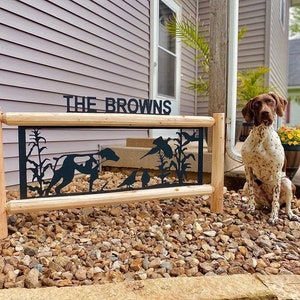 May include: A black metal sign with the text "THE BROWNS" and a silhouette of a dog and birds in a field of corn. The sign is mounted on two wooden posts. A brown and white dog is sitting in front of the sign.