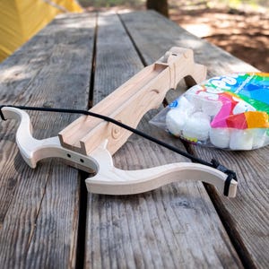 May include: A wooden toy crossbow with a black string, resting on a weathered wooden picnic table. A clear plastic bag filled with white foam balls and colorful packaging sits nearby. A yellow tent is visible in the background.