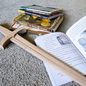 Puede incluir: Una espada de juguete de madera descansa sobre una alfombra junto a una pila de libros, incluyendo "The Hero and the Crown". Un libro abierto muestra ilustraciones y texto. La espada es de color marrón claro.