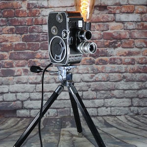 May include: A vintage Bolex movie camera repurposed as a lamp, featuring a black and silver metal body mounted on a three-legged tripod. An Edison-style light bulb with a glowing filament is attached to the top, emitting a warm light.