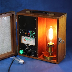 May include: A vintage wooden box with an open front revealing a glowing Edison bulb lamp. The box has a hinged door with a document inside. A control panel with gauges and dials is visible. A small light bulb is on the table.