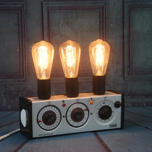 May include: A white vintage-style lamp with three light bulbs and three round dials. The lamp has a black base and a white cord.