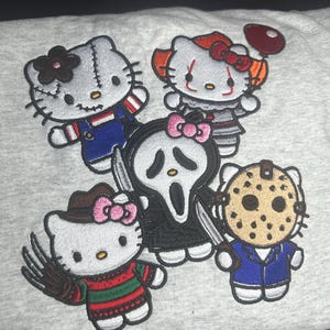 May include: A light grey sweatshirt featuring five embroidered Hello Kitty characters dressed as horror movie icons, including Chucky, Pennywise, Ghostface, Freddy Krueger, and Jason Voorhees, each with detailed colourful outfits and accessories.