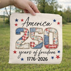 Pode incluir: Uma almofada creme com a inscrição "America 250 years of freedom 1776-2026". O número "250" é feito de tecido de retalhos em padrões vermelhos, brancos e azuis, adornado com estrelas vermelhas e azuis.