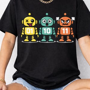 Puede incluir: Camiseta negra con tres robots de dibujos animados en amarillo, turquesa y naranja. Cada robot tiene una expresión diferente y un número: 01, 10 y 11. Los robots están sobre una línea blanca.