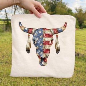 May include: Cream tote bag with a bull skull design. The skull is decorated with the American flag, featuring blue stars and red and white stripes. Two feathers hang from the horns. The bag is held by a hand.