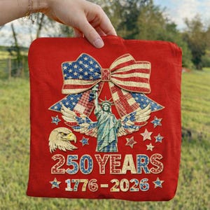 Pode incluir: Têxtil quadrado vermelho com um design patriótico. Apresenta um grande laço com o padrão da bandeira americana, a Estátua da Liberdade, uma águia, estrelas e o texto "250 YEARS 1776-2026".