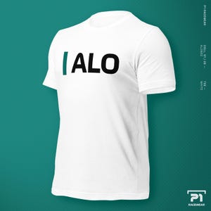 K&ouml;nnte beinhalten: Wei&szlig;es T-Shirt mit Rundhalsausschnitt und dem Text "|ALO" in Schwarz und T&uuml;rkis. Das Shirt hat kurze &Auml;rmel und wird vor einem t&uuml;rkisfarbenen Hintergrund pr&auml;sentiert. Der Text "P1 RACEWEAR" und "COLL. S1/26 - ALONSO" sind seitlich aufgedruckt.