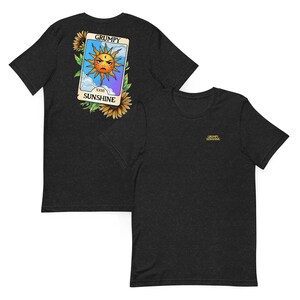 May include: Dark gray t-shirts. One shirt features a tarot card design with a grumpy sun, sunflowers, and the words "GRUMPY SUNSHINE." The other shirt has "GRUMPY SUNSHINE" embroidered on the chest.