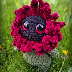 May include: A handmade crochet plush toy in the shape of a flower. The flower has a dark red, textured petal design, a black center with large, reflective eyes, and a green stem. The toy is set in a grassy environment.