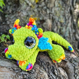 May include: A handmade green crochet dragon with rainbow accents on its horns, feet, and wings. The dragon has large, shiny black eyes and is lying on a textured, brown surface.