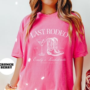May include: Pink oversized t-shirt with white text that reads "Last Rodeo" with a martini glass and cowboy boots graphic. The text also includes "June 2024, Emily's Bachelorette, Nashville, TN."