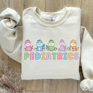 Peut inclure: Un sweat-shirt beige clair avec un motif de cinq dinosaures color&eacute;s et le mot "PEDIATRICS" en lettres arc-en-ciel.