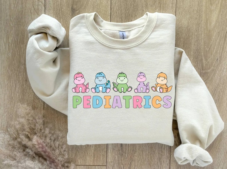 Peut inclure: Un sweat-shirt beige clair avec un motif de cinq dinosaures color&eacute;s et le mot "PEDIATRICS" en lettres arc-en-ciel.