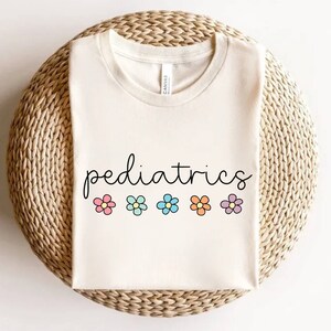 May include: Cream-colored t-shirt with the word "pediatrics" in a whimsical script font. Below the word are five colorful flower graphics in pink, light blue, blue, orange, and purple. The shirt is displayed on a woven, circular surface.