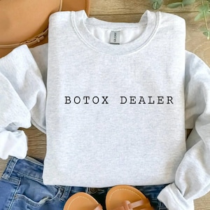 May include: A light gray sweatshirt with the text "BOTX DEALER" in black. The sweatshirt is displayed with blue jeans, a brown hat, and sandals.