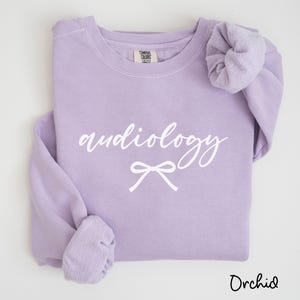May include: A light purple sweatshirt with the word "audiology" in white script and a white bow graphic. The word "Orchid" is written in a script font at the bottom right corner. The sweatshirt has long sleeves.