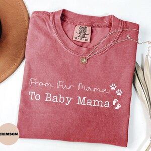 May include: Crimson-colored t-shirt with the text "From Fur Mama To Baby Mama" in white script. The shirt features paw prints and baby footprints. A silver necklace with a pendant rests on the shirt.
