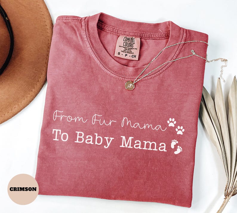 May include: Crimson-colored t-shirt with the text "From Fur Mama To Baby Mama" in white script. The shirt features paw prints and baby footprints. A silver necklace with a pendant rests on the shirt.