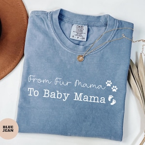May include: A blue jean colored t-shirt with the text "From Fur Mama To Baby Mama" in white script. The shirt has paw prints and baby footprints. A gold necklace with a pendant rests on the shirt.