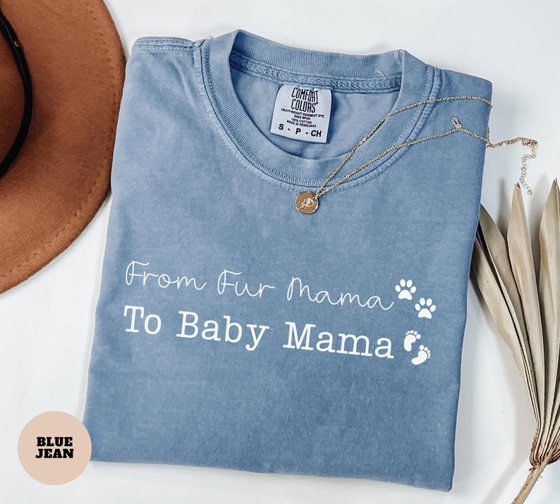 May include: A blue jean colored t-shirt with the text "From Fur Mama To Baby Mama" in white script. The shirt has paw prints and baby footprints. A gold necklace with a pendant rests on the shirt.