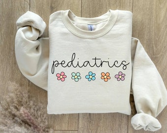 May include: Cream-colored sweatshirt with the word "pediatrics" in a handwritten font. Below the word are five colorful flower graphics in pink, blue, and orange. The sweatshirt has long sleeves and a crew neck.